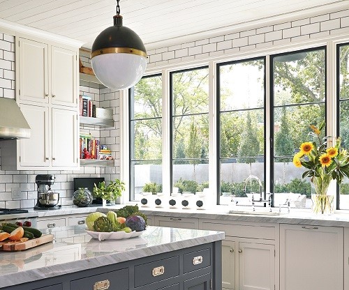 A white kitchen with white subway tiling on the walls, white cabinets, a granite-topped island, and a wall of large windows.